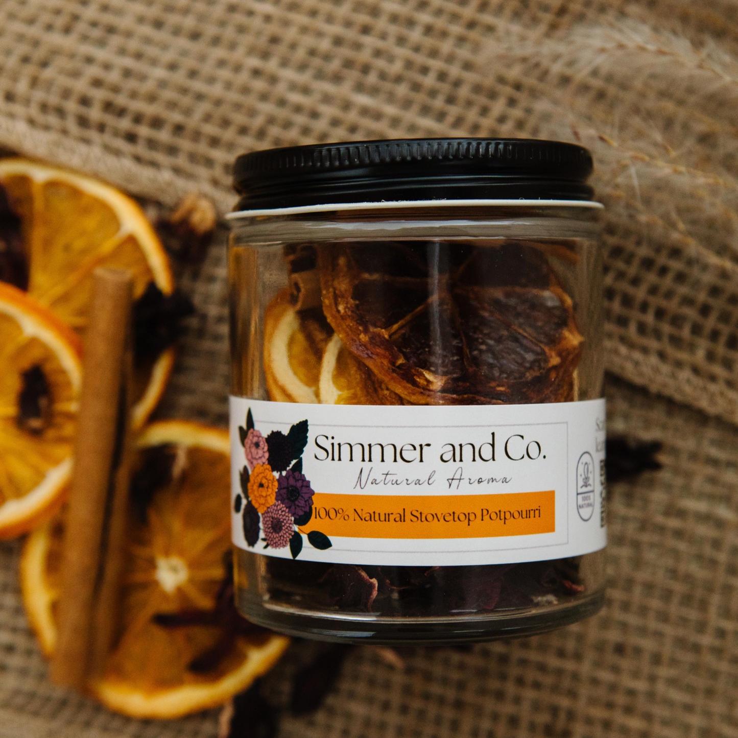 Jar of 'Simmer and Co.' natural stovetop potpourri with dried oranges and cinnamon sticks on a burlap background.